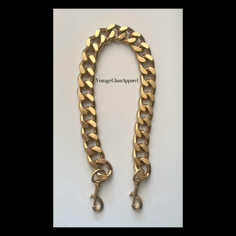 May include: A gold chain handbag strap with two clasps. The chain is thick and has a curb link design.