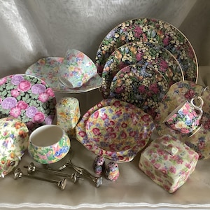 Peut inclure: Un ensemble de porcelaines à motifs floraux, comprenant des assiettes, des tasses, des soucoupes, un sucrier et une boîte couverte. La porcelaine présente divers motifs floraux roses, violets, jaunes et verts sur fond noir et crème.