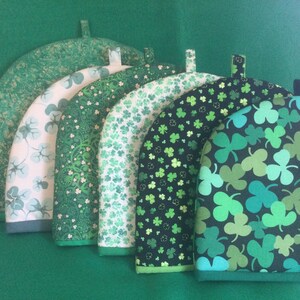 May include: Six green tea cozy covers with different shamrock patterns. The covers are made of fabric and have a loop for hanging.