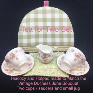 May include: A green and white checkered tea cozy with the text "Tea for Two Set" and a matching hotpad. Two white teacups with pink floral designs and saucers, and a small white creamer with a pink floral design are on a green floral fabric. The text "Teacozy and Hotpad made to Match the Vintage Duchess June Bouquet Two cups / saucers and small jug" is below the teacups.