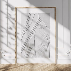 Peut inclure: Une impression d'art abstrait encadrée avec un motif texturé blanc et gris. L'impression est dans un cadre en bois clair et est accrochée à un mur blanc.