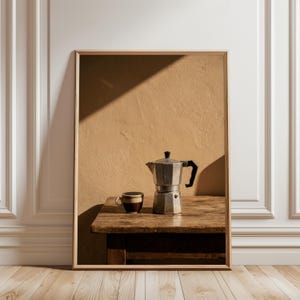 Puede incluir: Fotografía enmarcada con una naturaleza muerta de café. Una cafetera italiana plateada y una pequeña taza de café a rayas marrones y blancas están sobre una mesa de madera rústica. El fondo es una pared beige con una sombra.