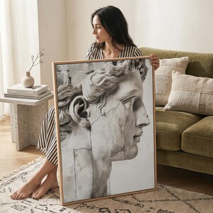 May include: A framed black and white print of a classical sculpture's profile. The artwork features a detailed depiction of a marble bust with visible cracks, set within a light wooden frame. The print is held in front of a green sofa.