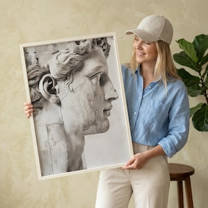 May include: A framed black and white print of a classical sculpture's profile. The artwork features a detailed depiction of a male face with visible cracks, set against a white background. The frame is a light beige color.
