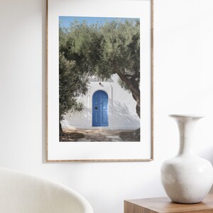 Peut inclure: Impression encadr&eacute;e d'une porte bleue dans un mur blanc, avec des arbres de chaque c&ocirc;t&eacute;. La porte est arqu&eacute;e et comporte deux panneaux. L'impression est dans un cadre en bois clair et est expos&eacute;e sur un mur blanc. Un vase blanc et une table en bois sont au premier plan.