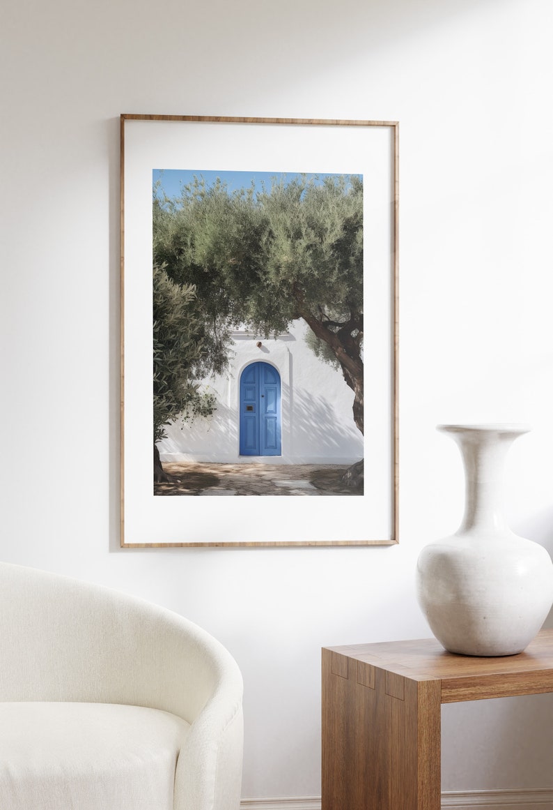 Peut inclure: Impression encadr&eacute;e d'une porte bleue dans un mur blanc, avec des arbres de chaque c&ocirc;t&eacute;. La porte est arqu&eacute;e et comporte deux panneaux. L'impression est dans un cadre en bois clair et est expos&eacute;e sur un mur blanc. Un vase blanc et une table en bois sont au premier plan.