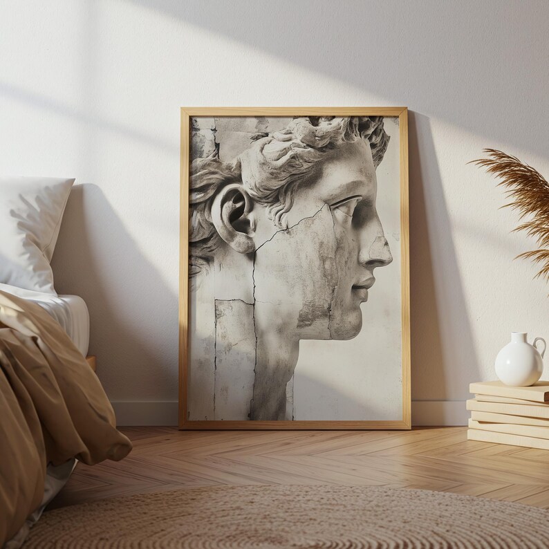 May include: A framed print featuring a grayscale depiction of a classical sculpture's head in profile. The artwork showcases a weathered appearance with visible cracks, set within a light wooden frame. The image is displayed against a neutral wall.