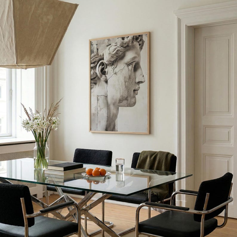 May include: A framed black and white print of a classical sculpture of a man's head hangs on a white wall. The room features a glass table with black chairs, a vase of flowers, and a modern light fixture.