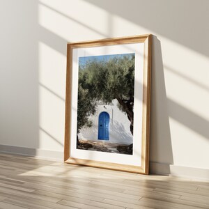 Peut inclure: Une impression encadr&eacute;e d'une porte bleue dans un mur blanc, sous un arbre vert. L'impression est dans un cadre en bois clair, appuy&eacute;e contre un mur blanc avec des ombres sur un sol en bois. L'image &eacute;voque la M&eacute;diterran&eacute;e.