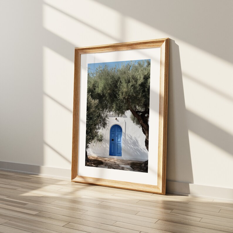 Peut inclure: Une impression encadr&eacute;e d'une porte bleue dans un mur blanc, sous un arbre vert. L'impression est dans un cadre en bois clair, appuy&eacute;e contre un mur blanc avec des ombres sur un sol en bois. L'image &eacute;voque la M&eacute;diterran&eacute;e.