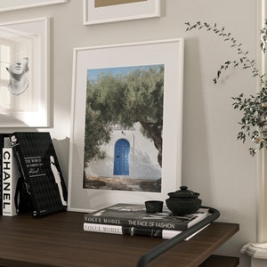 Peut inclure: Une impression encadr&eacute;e d'un b&acirc;timent blanc avec une porte bleue, un olivier et un ciel bleu. L'impression est sur une surface en bois fonc&eacute; avec des livres, un petit bol noir et un pot d&eacute;coratif. D'autres &oelig;uvres d'art encadr&eacute;es sont visibles.