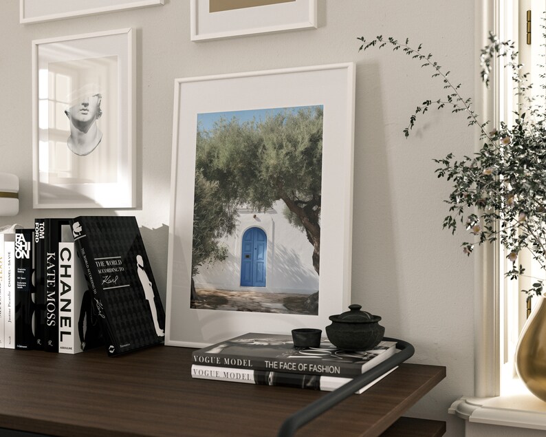 Peut inclure: Une impression encadr&eacute;e d'un b&acirc;timent blanc avec une porte bleue, un olivier et un ciel bleu. L'impression est sur une surface en bois fonc&eacute; avec des livres, un petit bol noir et un pot d&eacute;coratif. D'autres &oelig;uvres d'art encadr&eacute;es sont visibles.