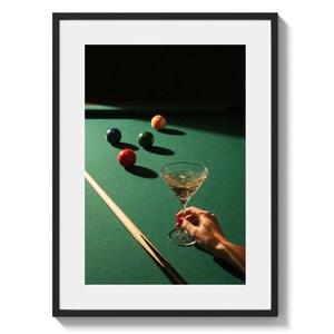 May include: A framed image of a pool table with billiard balls and a martini glass. The balls are red, blue, green, and orange. A cue stick rests on the green felt. A hand holds the martini glass.