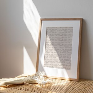 May include: A framed print with the words "AMOR PATI" repeated in a vertical pattern on a white background. The frame is light brown wood.