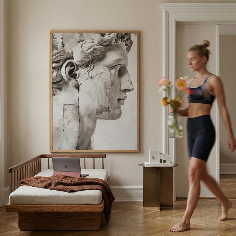 May include: A large framed print of a classical bust in shades of gray and white hangs above a wooden chaise lounge with a laptop and a brown blanket. A woman walks past holding flowers.