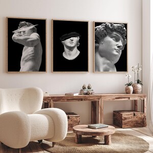 K&ouml;nnte beinhalten: Drei Schwarzwei&szlig;-Bilderrahmen mit Skulpturen. Die Bilder h&auml;ngen an einer wei&szlig;en Wand &uuml;ber einem Holztisch. Die Skulpturen zeigen einen M&auml;nnerkopf, einen M&auml;nnertorso und einen M&auml;nnerkopf mit Hut.