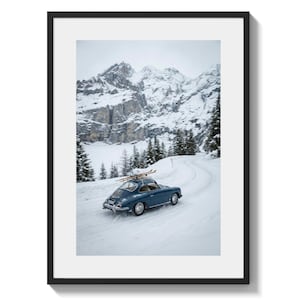 Puede incluir: Fotografía enmarcada de un coche azul vintage con esquís en el techo, conduciendo por una carretera nevada. El coche se encuentra frente a un telón de fondo de montañas cubiertas de nieve y árboles de hoja perenne, creando una escena invernal.