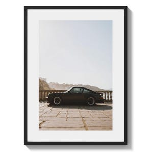 May include: Framed print of a black sports car parked on a stone patio with a scenic backdrop. The car has a sleek design with black wheels and a low profile. The print is in a black frame.