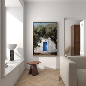 Peut inclure: Une impression encadr&eacute;e d'un b&acirc;timent blanc avec une porte bleu vif, partiellement cach&eacute;e par un grand olivier. L'impression, dans un cadre dor&eacute;, est expos&eacute;e dans une pi&egrave;ce moderne et lumineuse avec une fen&ecirc;tre et un tabouret en bois.