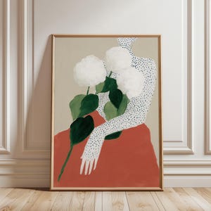 Peut inclure: Impression d'art encadrée représentant une figure tenant des fleurs blanches avec des feuilles vertes. Le bras et l'épaule de la figure sont recouverts de points noirs. La figure porte un vêtement rouge, sur fond beige.