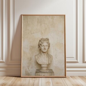 Puede incluir: Una lámina de arte enmarcada que presenta un fondo de pared de yeso desgastado en tonos beige, con un busto de piedra clara de una figura clásica con cabello rizado, colocado sobre una base de pedestal a juego dentro de un marco de madera.