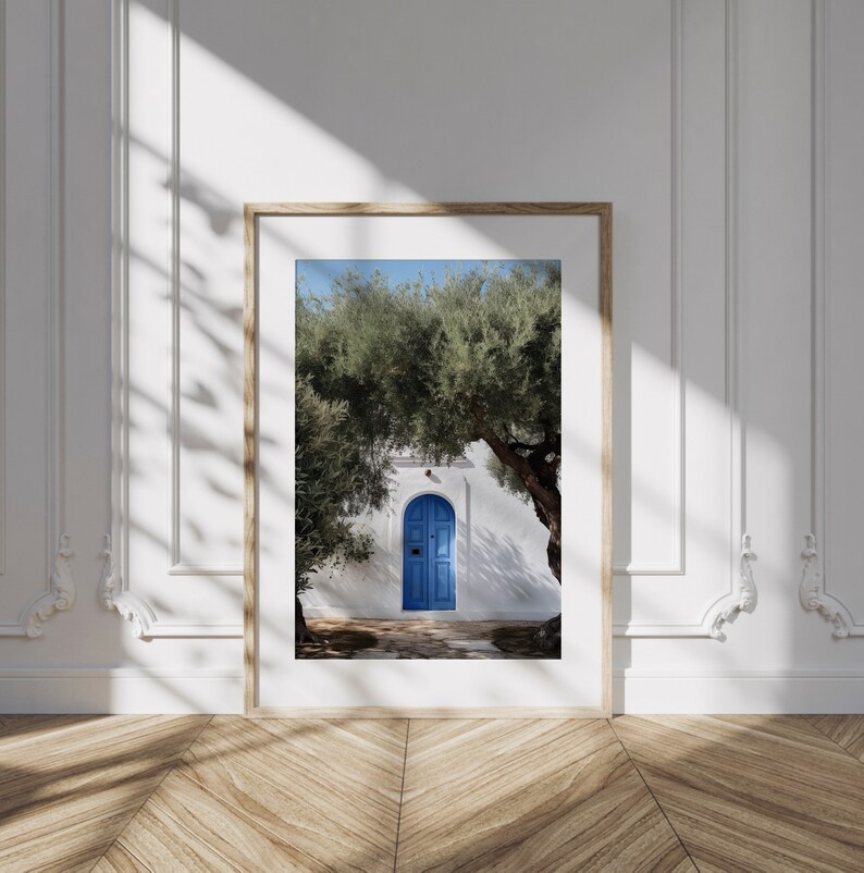 Peut inclure: Une impression encadr&eacute;e repr&eacute;sentant une porte bleue vive dans un b&acirc;timent blanc, encadr&eacute;e par des arbres verts. L'image est plac&eacute;e sur un ciel bleu clair, avec la lumi&egrave;re naturelle et les ombres ajoutant de la profondeur. Le cadre est de couleur bois clair.