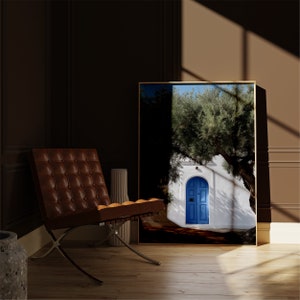 Peut inclure: Une impression d'art encadr&eacute;e repr&eacute;sentant un b&acirc;timent blanc avec une porte bleue, un grand arbre et un ciel bleu. L'impression est expos&eacute;e dans une pi&egrave;ce avec une chaise en cuir marron et un vase d&eacute;coratif. L'&oelig;uvre a un cadre dor&eacute;.