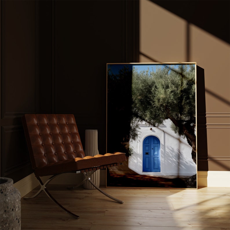 Peut inclure: Une impression d'art encadr&eacute;e repr&eacute;sentant un b&acirc;timent blanc avec une porte bleue, un grand arbre et un ciel bleu. L'impression est expos&eacute;e dans une pi&egrave;ce avec une chaise en cuir marron et un vase d&eacute;coratif. L'&oelig;uvre a un cadre dor&eacute;.