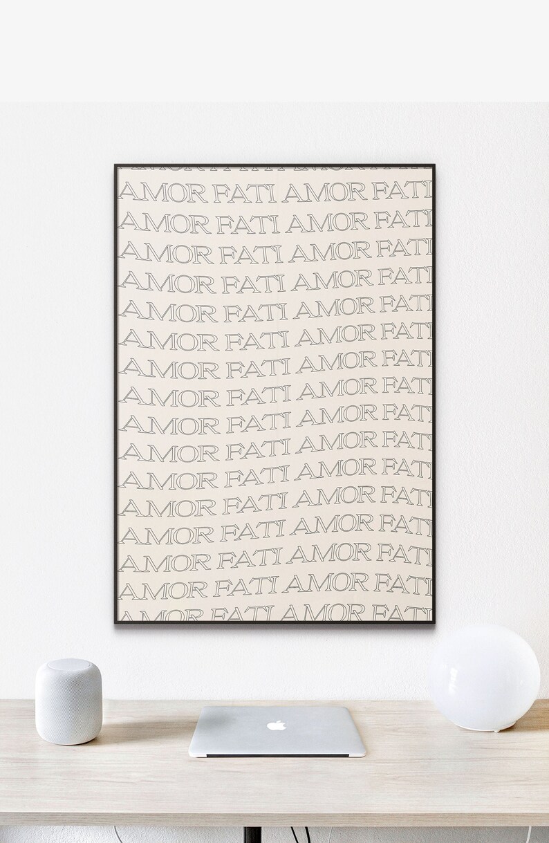 May include: A framed print with the phrase "Amor Fati" repeated in a minimalist style, printed in black and white on a cream background. The print is hanging on a white wall above a desk with a laptop and two decorative objects.