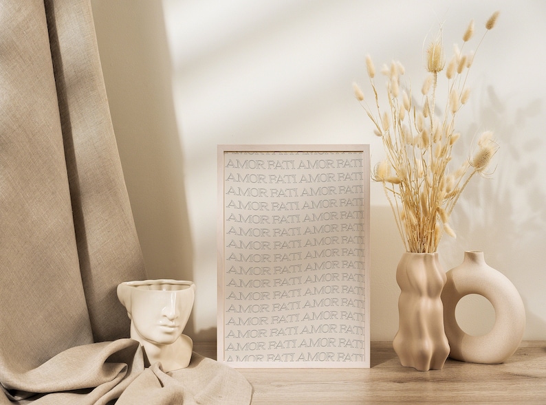 May include: A framed print with the text "AMOR PATI" repeated in a beige color on a white background. The print is sitting on a wooden table with a beige vase with dried flowers and a white ceramic vase with a face design.