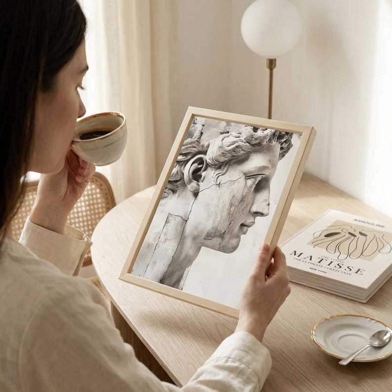 May include: A framed black and white print of a classical sculpture's profile. The print is held by a person, with a book titled "MATISSE" visible nearby. A cup of coffee and a saucer with a spoon are on the table.