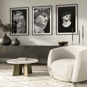 K&ouml;nnte beinhalten: Drei schwarz gerahmte Drucke von Schwarzwei&szlig;fotos von Skulpturen. Die Drucke h&auml;ngen an einer wei&szlig;en Wand &uuml;ber einem wei&szlig;en Sessel und einem wei&szlig;en Couchtisch.