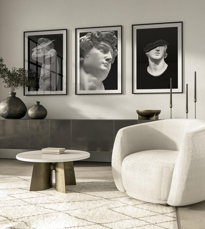 K&ouml;nnte beinhalten: Drei schwarz gerahmte Drucke von Schwarzwei&szlig;fotos von Skulpturen. Die Drucke h&auml;ngen an einer wei&szlig;en Wand &uuml;ber einem wei&szlig;en Sessel und einem wei&szlig;en Couchtisch.
