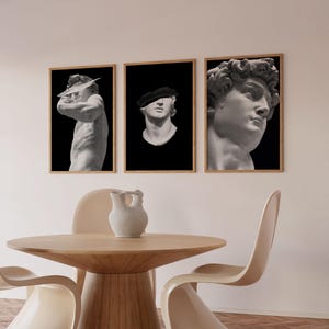 K&ouml;nnte beinhalten: Drei gerahmte Schwarzwei&szlig;drucke von klassischen Skulpturen. Die Drucke h&auml;ngen an einer wei&szlig;en Wand &uuml;ber einem Holztisch mit zwei beigen St&uuml;hlen.