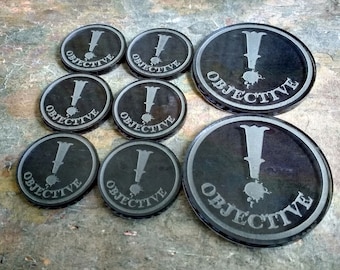 May include: Six black round tokens with a white exclamation point and the word "OBJECTIVE" printed on each. The tokens are arranged in a circle on a textured surface.