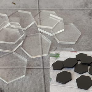 May include: Clear, hexagonal acrylic game tokens are scattered across a textured surface. A smaller group of black hexagonal tokens is in the lower right corner. These tokens are likely used for board games or tabletop gaming.