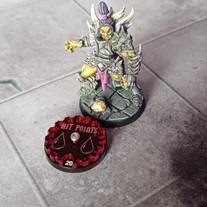 Puede incluir: Un contador de puntos de golpe de plástico rojo con un diseño de engranaje y dos formas de lágrima. El contador tiene las palabras "Hit Points" impresas y el número "20" impreso en la parte inferior.