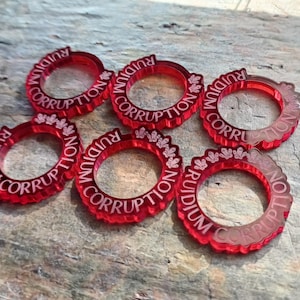 May include: Seven red plastic tokens with the words "RUIDIUM CORRUPTION" engraved in white. The tokens are circular and have a bevelled edge.