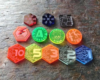 May include: A collection of colorful acrylic game tokens. The tokens are various shapes, including hexagons and circles, and feature designs such as circuit board patterns, numbers, and icons. Colors include red, orange, green, yellow, and blue. Some tokens have text like "CORE DAMAGE" and "BAD PUBLICITY".