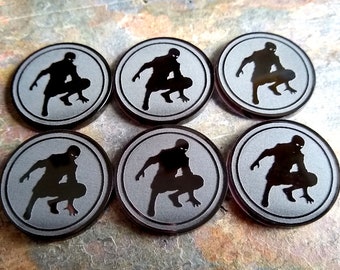 May include: Six circular tokens arranged in two rows of three. Each token features a matte grey surface with a black border and a central black silhouette of a crouching figure in a dynamic pose. The tokens have a smooth, flat design.