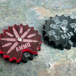 May include: Two red and black plastic gear-shaped tokens. The red token has a design of bullets and the text "AMMO 10". The black token has a design of pistons and a gear with the text "GEAR 6".