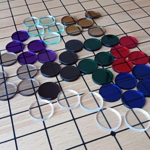 Puede incluir: Colección de fichas de juego redondas y coloridas en varios colores, incluyendo transparente, morado, negro, verde, rojo y azul. Las fichas están dispuestas sobre una superficie clara con un patrón de cuadrícula.