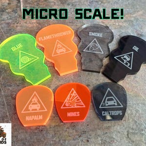 Puede incluir: Un conjunto de ocho fichas de peligro de acrílico para un juego de mesa. Las fichas tienen forma de triángulos con diferentes símbolos de peligro y texto, incluyendo "Napalm", "Minas", "Caltrops", "Aceite", "Humo", "Lanza llamas" y "Azul".