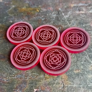 Puede incluir: Cinco fichas circulares rojas con un diseño geométrico blanco en el centro. El diseño es una serie de cuadrados y círculos concéntricos.