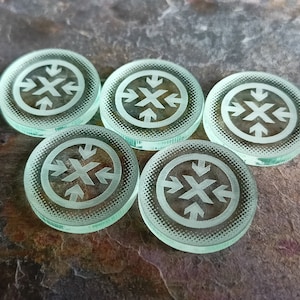May include: Five circular, pale green glass tokens featuring a central cross design with outward-pointing arrows. The tokens have a frosted finish and are arranged on a textured, dark brown surface.