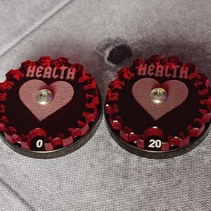 May include: Two red plastic health trackers with a heart design and numbers 0 to 20. The word "HEALTH" is printed on each tracker.