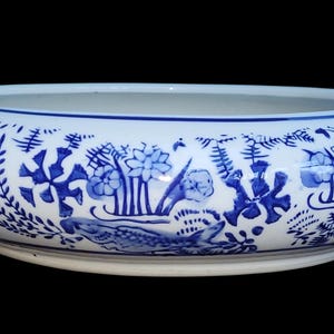 May include: A white ceramic bowl with a blue floral and foliage design. The bowl has a wide, shallow shape and a smooth, glossy finish. The blue design features stylized flowers, plants, and other decorative elements. The bowl is set against a black background.