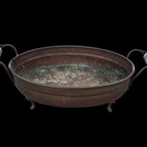 May include: An oval copper bowl with a weathered patina interior, featuring two dark metal handles adorned with white and blue floral ceramic accents. The bowl is supported by four small, curved metal feet, showcasing a rustic appearance.