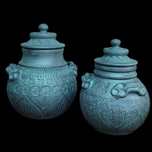 May include: Two teal ceramic jars with lids, featuring embossed patterns and decorative handles. The jars have a rounded shape and matching lids with a small knob. The background is black.