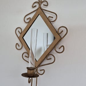 May include: A decorative wall mirror with a diamond-shaped frame and gold-coloured scrollwork. A clear glass candle holder is attached to the bottom. The mirror reflects the candle holder and the surrounding wall.